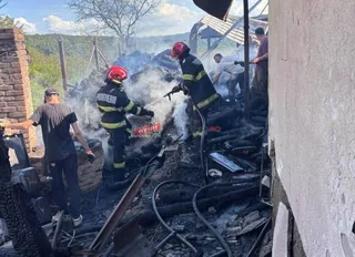 Pompieri în echipament de protecție și un bărbat stau lângă resturile carbonizate ale unei structuri de unde se ridică fum alb.