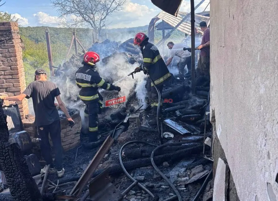 Pompieri în echipament de protecție și un bărbat stau lângă resturile carbonizate ale unei structuri de unde se ridică fum alb.