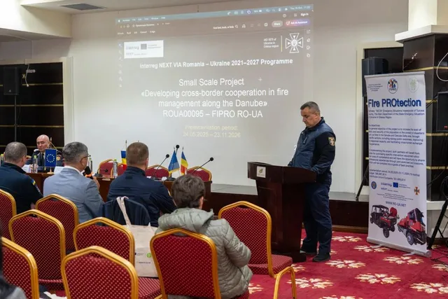 Un bărbat stă la un podium în fața unui ecran de proiecție, în timp ce un grup de persoane stă pe scaune în fața lui.