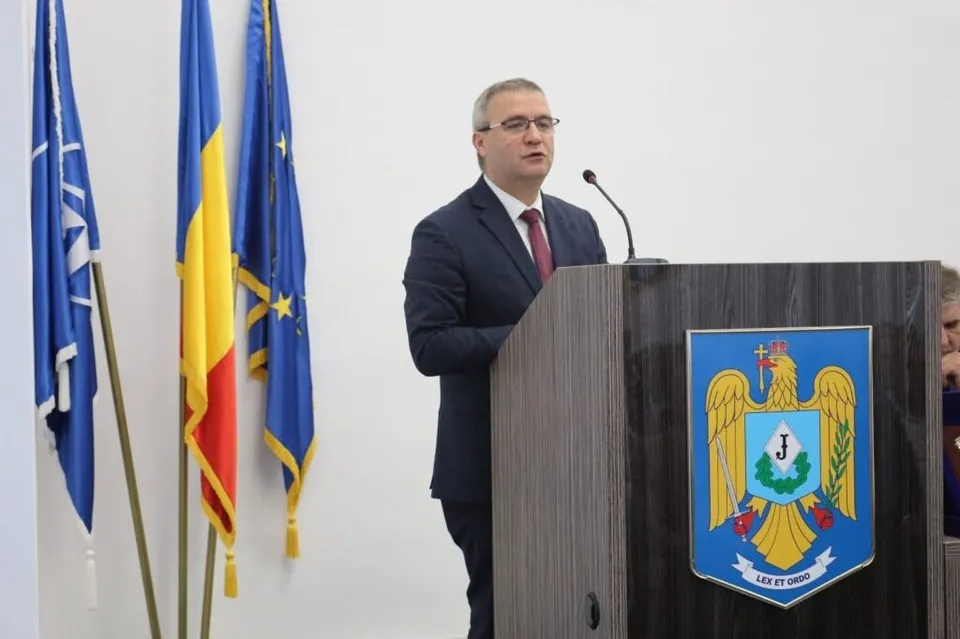Un bărbat cu ochelari, purtând costum închis și cravată roșie, stă în spatele unui podium lângă trei steaguri pe un fundal alb.