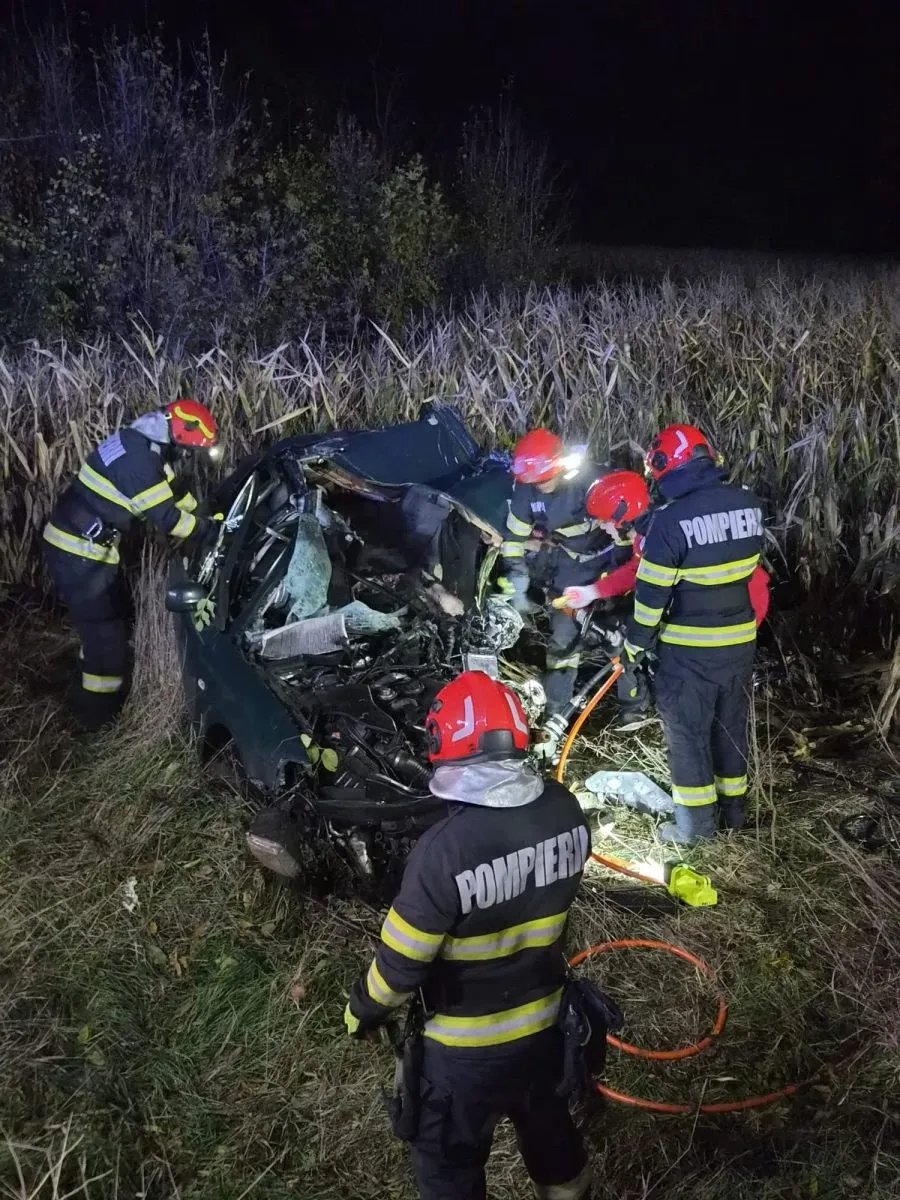 Pompierii în echipament complet de protecție cu helmeți roșii intervin noapta lângă o mașină accidentată complet zdrobită, pe un teren cu vegetație înalță.