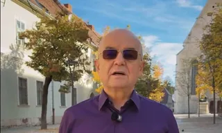 Un bărbat cu ochelari de soare și cămașă mov stă în fața unor clădiri și copaci sub un cer senin.