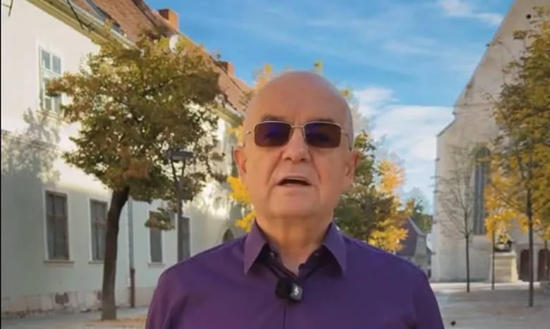 Un bărbat cu ochelari de soare și cămașă mov stă în fața unor clădiri și copaci sub un cer senin.