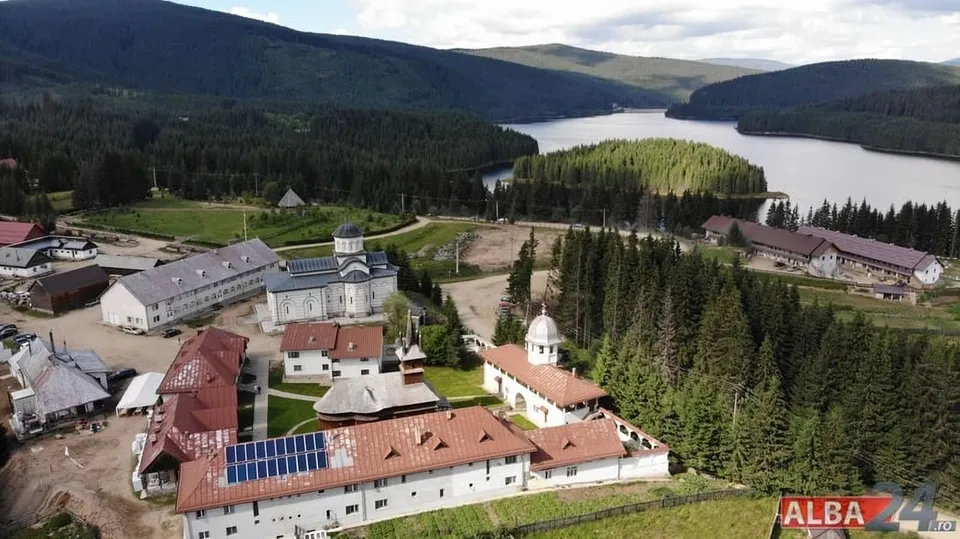 O vedere aeriană prezintă un complex de clădiri cu acoperișuri închise și o mănăstire situată într-o zonă montană cu păduri și un lac.