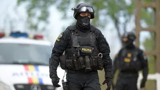 Un agent de poliție în uniformă neagră, cu vestă tactică și mască, merge pe o stradă lângă un vehicul de poliție alb.