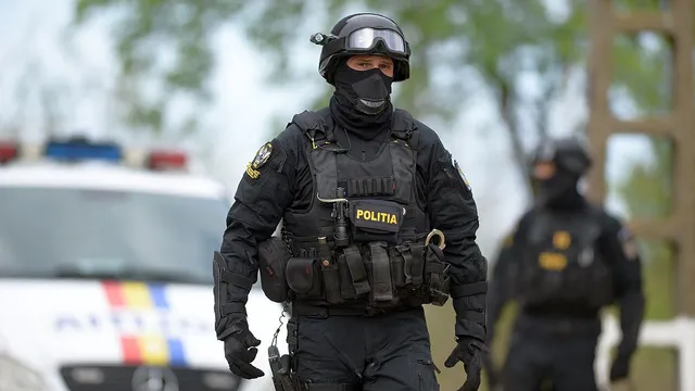 Un agent de poliție în uniformă neagră, cu vestă tactică și mască, merge pe o stradă lângă un vehicul de poliție alb.