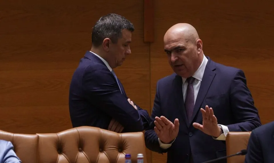 Un bărbat cu părul scurt și costum albastru stă în profil, în timp ce un bărbat chel cu costum închis și cămașă albă face gesturi cu mâinile.