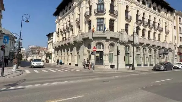 O stradă asfaltată cu marcaje rutiere traversează în fața unui clădire înaltă, de culoare deschisă, cu ferestre și balcoane întemeiate.