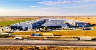 O vedere aeriană prezintă un complex de clădiri industriale gri cu acoperișuri plate, situate lângă o autostradă pe unde circulă mai multe camioane și mașini.