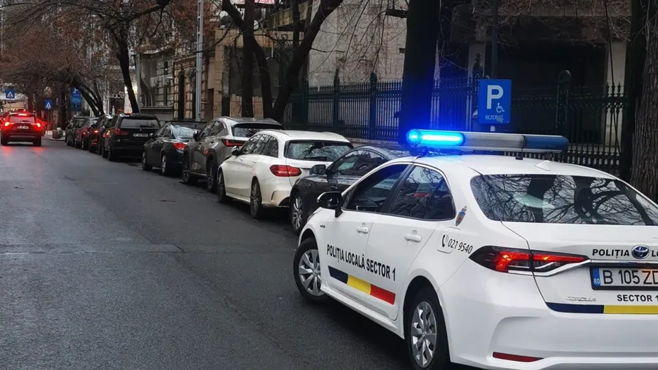 Un autovehicul alb cu inscripția Politia Locala Sector 1 este parcat pe marginea unui drum lângă alte mașini aliniate pe aceeași parte.