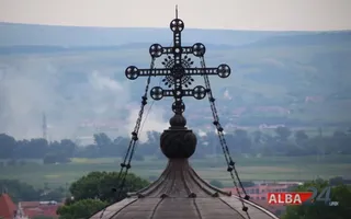 O cruce metalică decorată este montată pe vârful unei cupole, având un peisaj cu dealuri și vegetație în fundal.