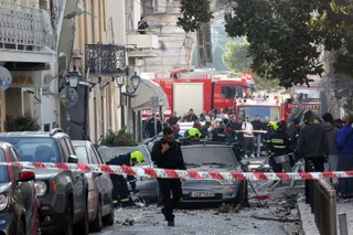 O mulțime de oameni și vehicule de intervenție roșii sunt situate pe un drum strâmt lângă mașini parcate și o zonă delimitată cu bandă de alertă.