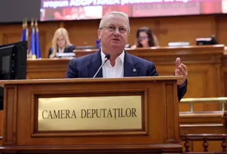 Un bărbat cu ochelari și costum închis stă în spatele unui podium de lemn pe care scrie Camera Deputaților.