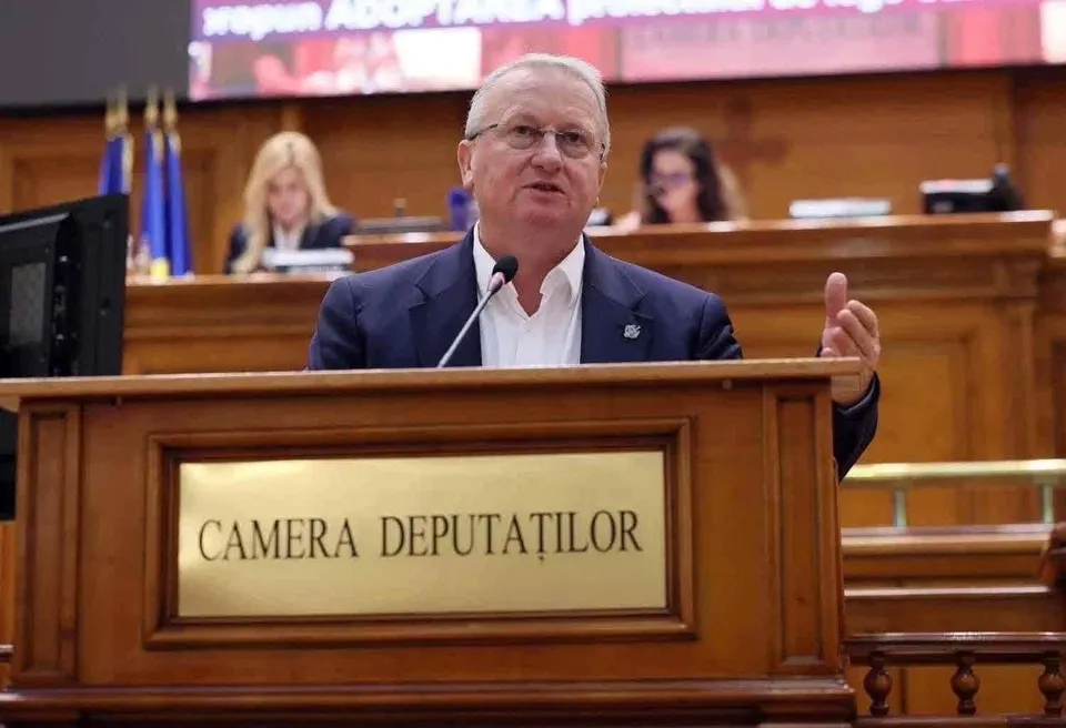 Un bărbat cu ochelari și costum închis stă în spatele unui podium de lemn pe care scrie Camera Deputaților.