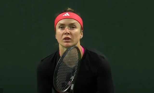 O femeie cu bandană roșie și tricou negru ține o raketă de tenis în fața unui fundal verde uniform.