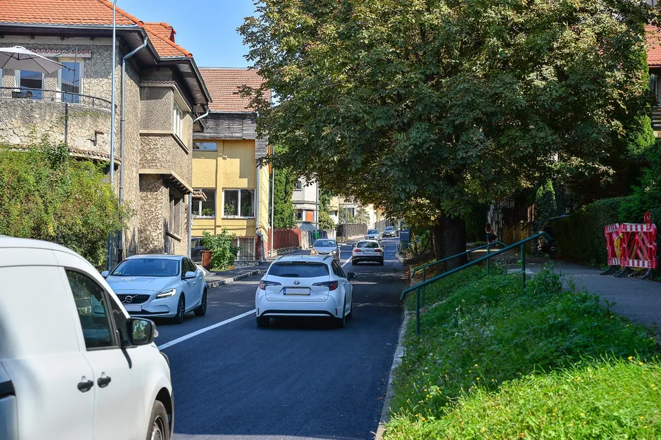 Mașini circulă pe un drum asfaltat recent, lângă clădiri și un copac mare, într-o zonă cu vegetație și un gard metalic verde.