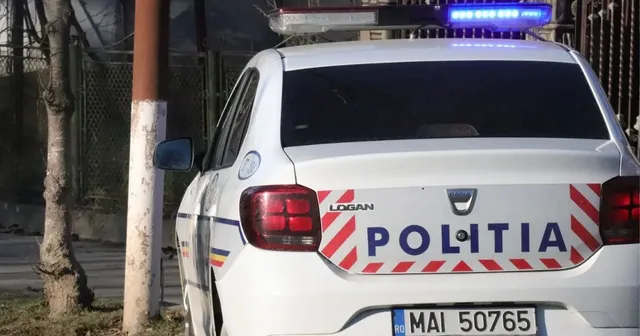 O mașină de poliție albă cu marcajul POLITIA este parcată pe marginea unui drum lângă un copac și un stâlp.