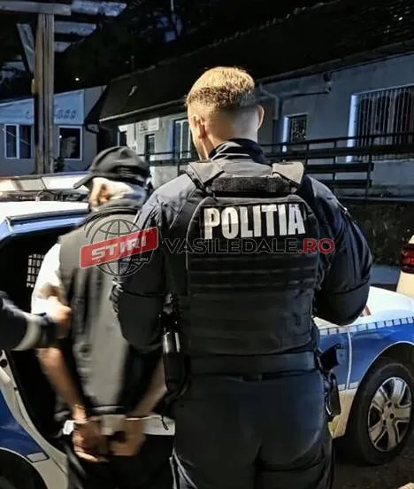 Un polițist cu un vest vest vest de protecție pe spate stă lângă un vehicul alb lângă un bărbat cu șapcă și vestă neagră.