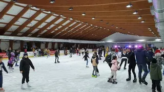 Oamenii patinează pe o suprafață de gheață sub un plafon înalt cu grinzi de lemn și lumini aliniate.