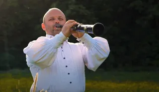 Un bărbat cu capul chel și cămașă albă cu mâneci largi suflă într-un instrument muzical lung de culoare închisă.