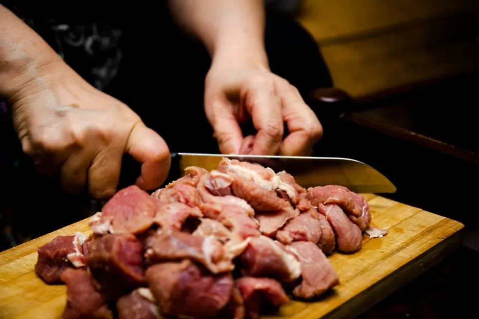 Mâinile unei persoane taie bucăți de carne crudă pe un tocător de lemn folosind un cuțit metalic.