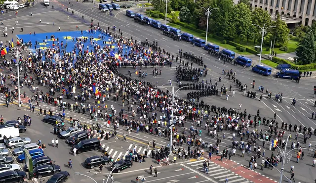 O mulțime de oameni protestează într-o piață, cu un steag mare al Uniunii Europene pe jos, înconjurați de forțe de ordine și vehicule de poliție. 
