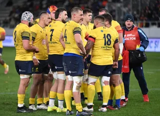 Un grup de sportivi în echipamente galbene și blu de rugby stau adunați pe un teren de sport verde.