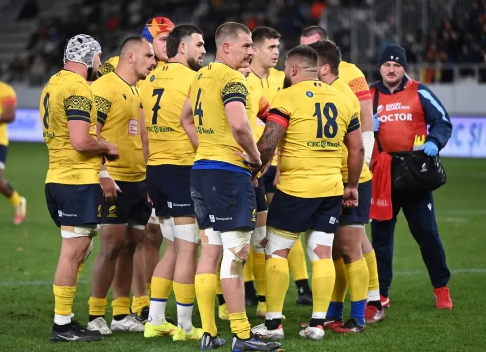 Un grup de sportivi în echipamente galbene și blu de rugby stau adunați pe un teren de sport verde.