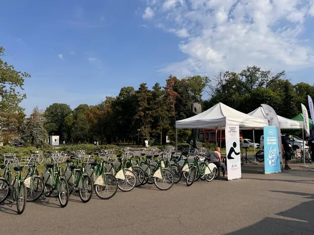 O serie de biciclete verzi sunt parcate în linie pe un teren asfaltat lângă două corturi albe sub un cer senin.