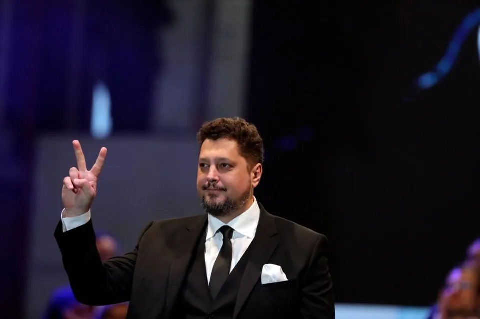 Un bărbat cu barbă, îmbrăcat într-un costum negru și cămașă albă, face semnul victoria cu mâna dreaptă.