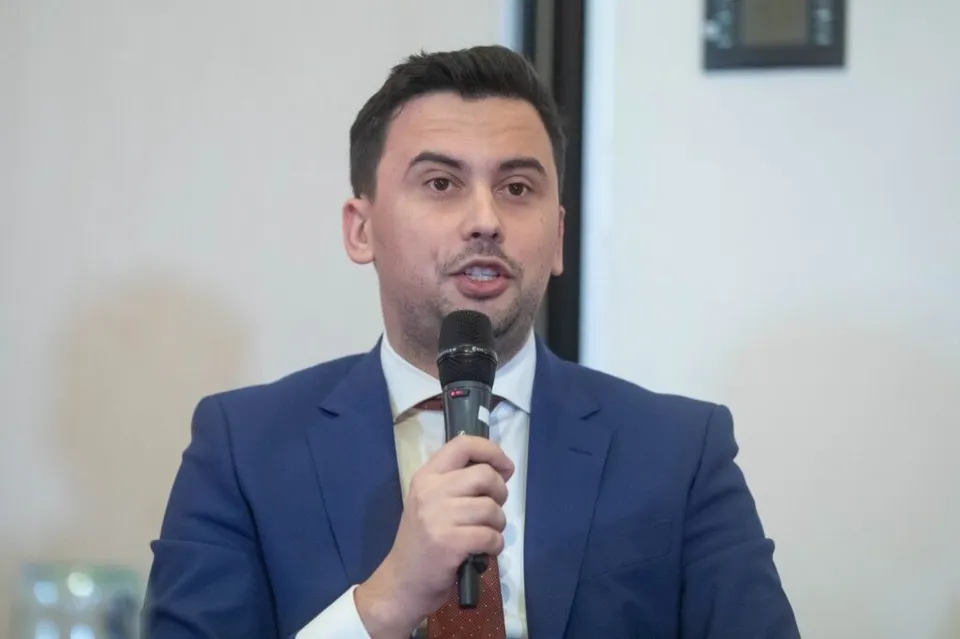 Un bărbat cu părul scurt, purtând sacou albastru, cămașă albă și cravată, vorbește într-un microfon negru ținut în mână.