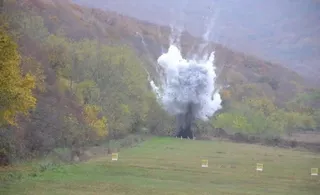 O explozie de culoare albă se ridică deasupra unui câmp verde lângă un versant de deal acoperit de vegetație autumnală.