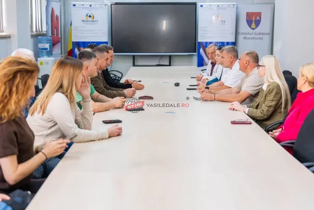 O grupă de persoane stă la o masă lungă și albă într-o cameră cu un ecran negru montat pe perete în fundal.