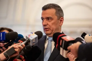 Un bărbat într-un costum închis la culoare și cămașă albă cu cravată deschisă vorbește în fața mai multor microfoane de presă.