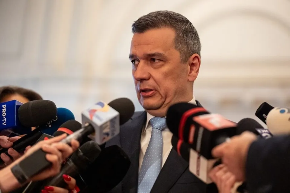 Un bărbat într-un costum închis și cămașă albă cu cravată deschise la culoare vorbește în fața mai multor microfoane.