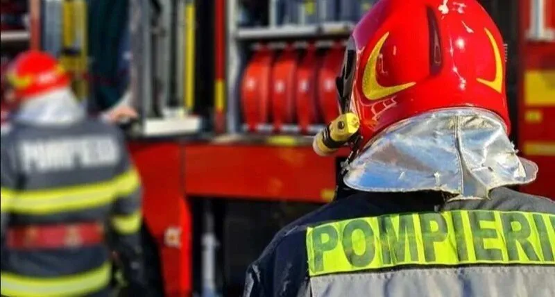 Un pompier cu cască roșie și echipament reflectorizant stă cu spatele, privind spre un vehicul de intervenție cu echipamente de stingere.