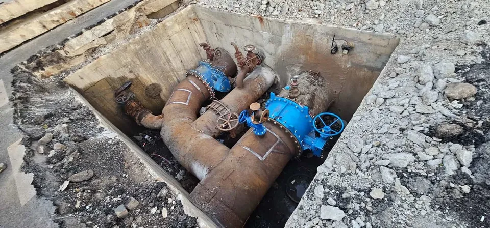 O conductă metalică mare, cu robinete și îmbinări albastre, este vizibilă într-o groapă deschisă în pământ.