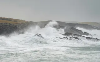 Valuri mari cu spumă albă se izbesc de pe un coaste de stânci sub un cer gri și înnorat.