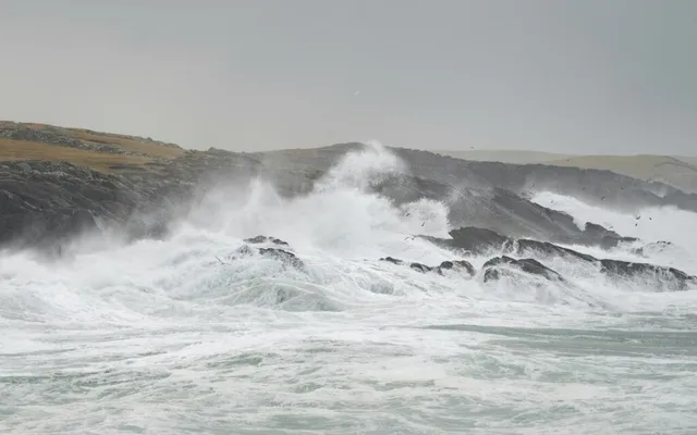 Valuri mari cu spumă albă se izbesc de pe un coaste de stânci sub un cer gri și înnorat.
