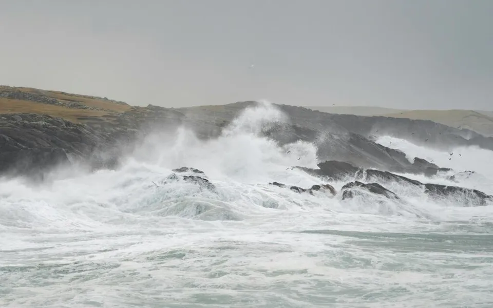 Valuri mari cu spumă albă se izbesc de pe un coaste de stânci sub un cer gri și înnorat.