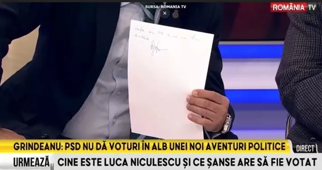 Un bărbat într-un costum închis și cămașă deschisă ține o foaie de hârtie albă cu o semnătură scrisă cu pixul.