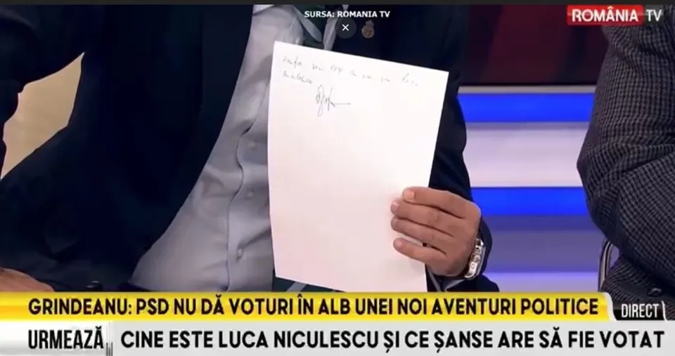 Un bărbat într-un costum închis și cămașă deschisă ține o foaie de hârtie albă cu o semnătură scrisă cu pixul.