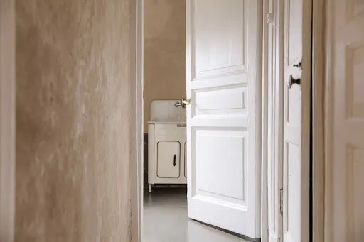 O ușă albă deschisă oferă o vedere spre un spațiu interior cu un aparat de spălat și un perete de culoare neutră.