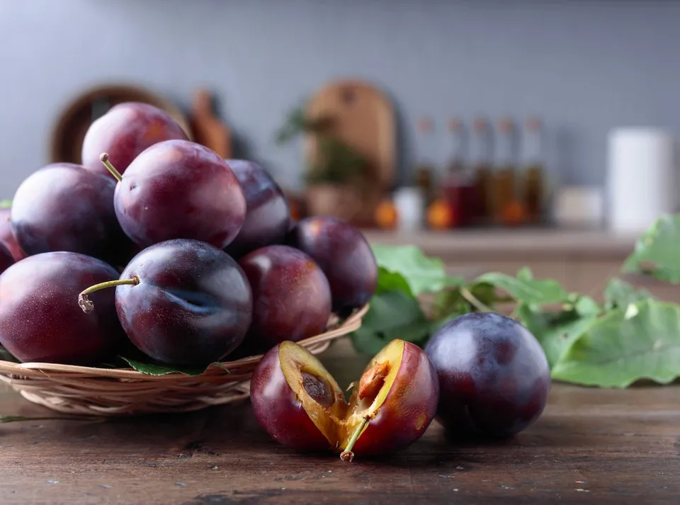 Prune mov închis într-un coș și pe o masă, cu o fructă tăiată și frunze verzi.