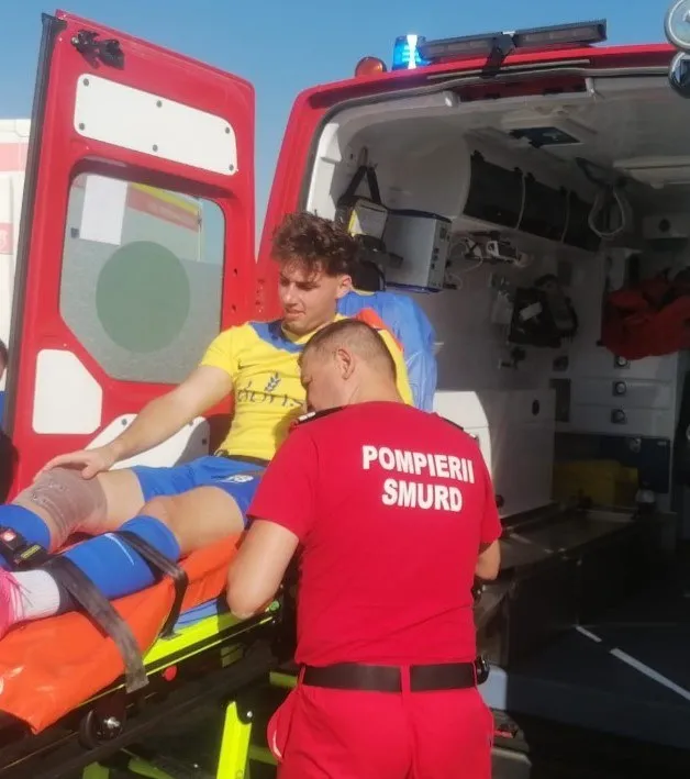 Un bărbat cu tricou galben stă întins pe o targă, fiind asistat de un salvator cu tricou roșu pe fundalul unei ambulanțe deschise.