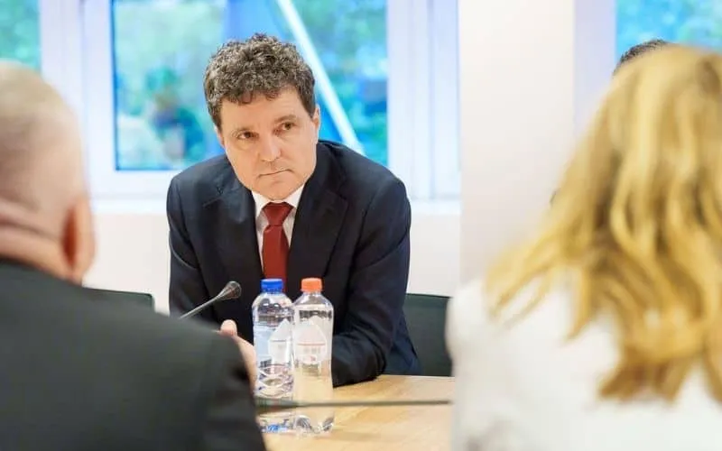 Un bărbat cu părul creț, purtând sacou închis și cravată roșie, stă la o masă lângă două sticle de apă, privind spre persoane din prim-plan.