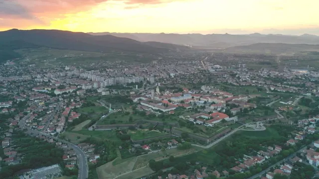 O vedere aeriană a unui oraș cu clădiri și fortificații, situat într-o vale cu dealuri și munți în fundal sub un cer la apus.