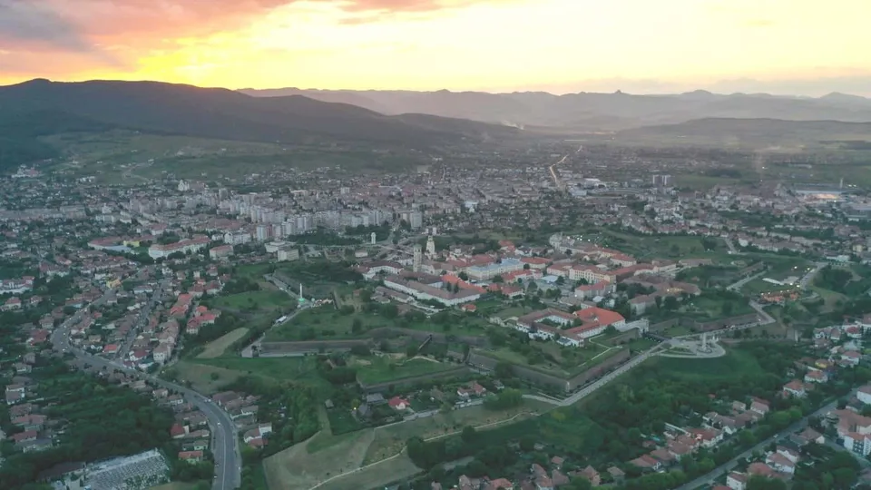 O vedere aeriană a unui oraș cu clădiri și fortificații, situat într-o vale cu dealuri și munți în fundal sub un cer la apus.