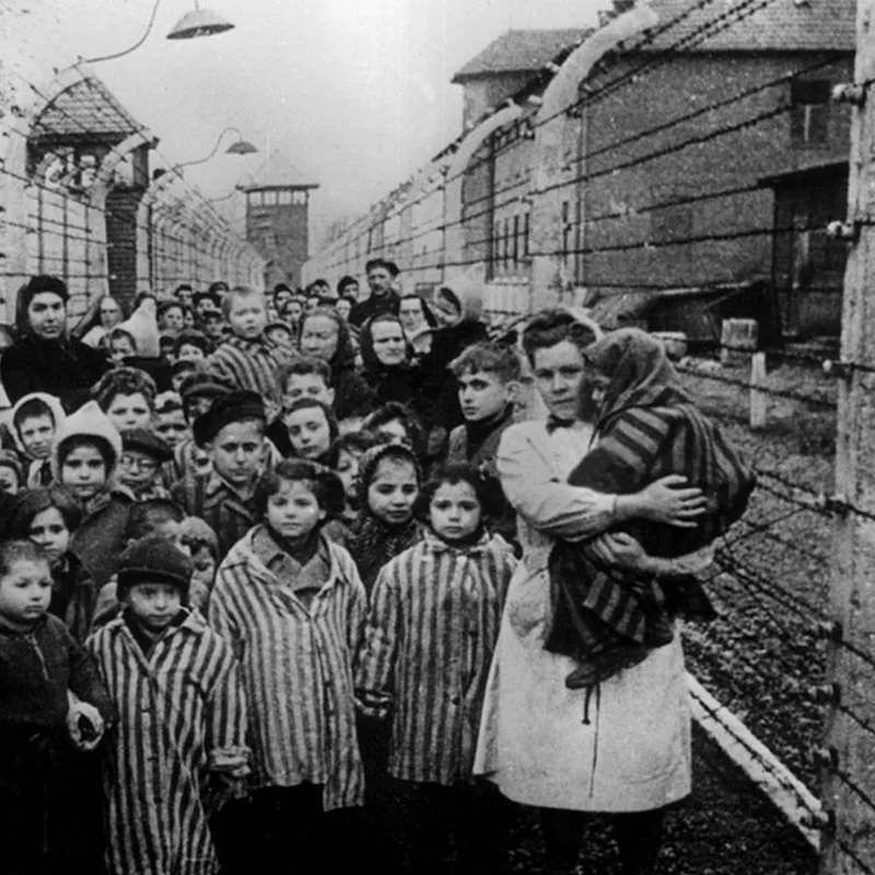 Un grup de copii și adulți îmbrăcați în haine dungate stau lângă un gard de sârmă ghimpată lângă niște clădiri și turnuri de supraveghere.