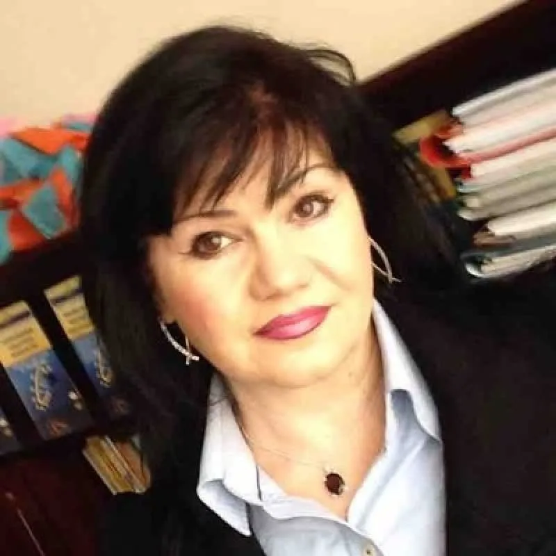 O femeie cu părul negru, purtând o cămașă deschisă la guler și un colier cu pandantiv, este pozată în fața unor dosare pe un raft.